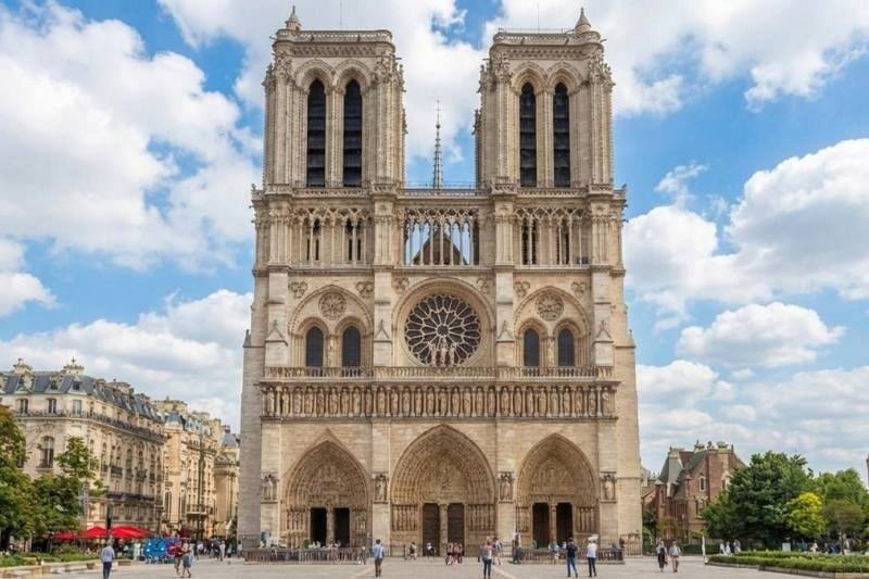 Front facade of Notre-Dame Cathedral in Paris showing its twin Gothic towers, rose window, and three arched portals under a partly cloudy sky