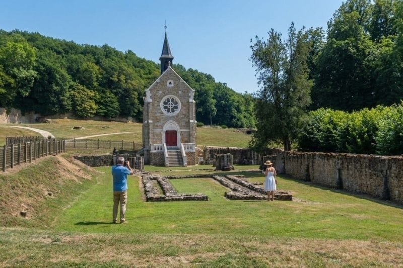 Two visitors photographing the ruins and small stone chapel at the historic Abbey of Port-Royal-des-Champs surrounded by green hills