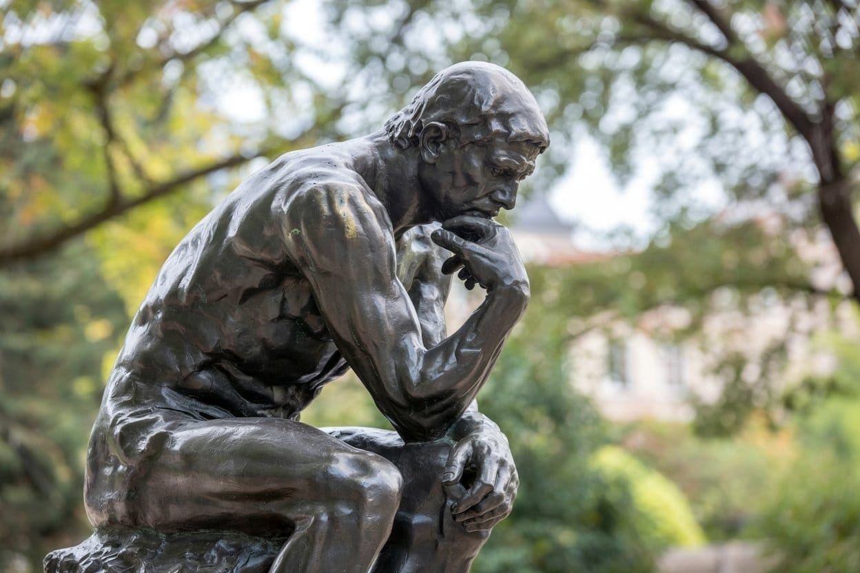 Close-up of The Thinker bronze sculpture by Auguste Rodin, seated with chin resting on hand, surrounded by autumn foliage